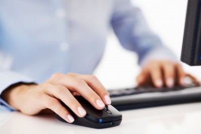 A computer user sat at a desk with one hand on a mouse another on the keyboard.