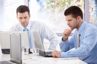Two men in shirts and ties sat a desk working with their laptops