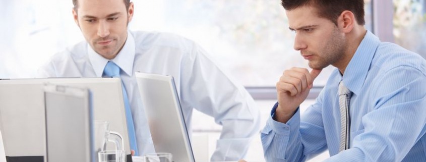 Two men in shirts and ties sat a desk working with their laptops